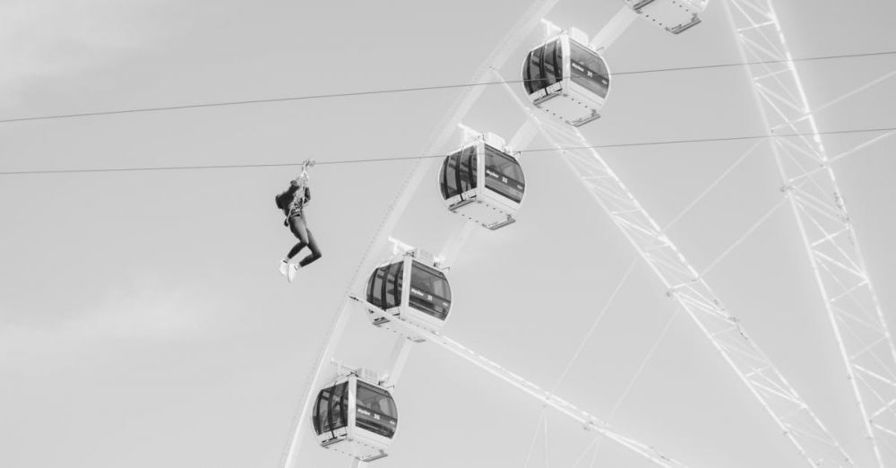 Thrilling Zip Lining - Thrilling black and white shot of a person zip lining past a Ferris wheel in a daring amusement park scene.