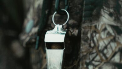 Safety In Fishing & Hunting - Close-up of a shiny silver whistle hanging against a camouflage-patterned fabric background.