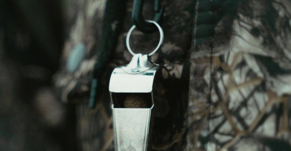 Safety In Fishing & Hunting - Close-up of a shiny silver whistle hanging against a camouflage-patterned fabric background.