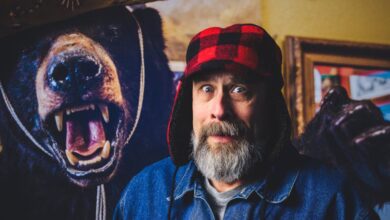 Bear Hunting - A bearded man looking surprised beside a bear mount in a cozy indoor environment.