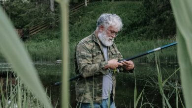 Solo Hunting - A senior man with grey hair fishes by a tranquil lake near a cabin surrounded by lush greenery.