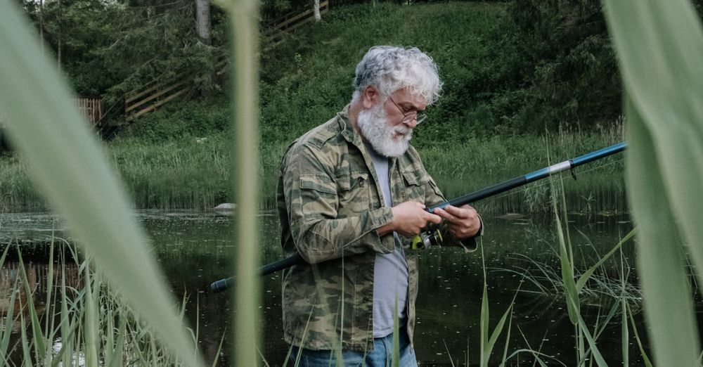 Solo Hunting - A senior man with grey hair fishes by a tranquil lake near a cabin surrounded by lush greenery.