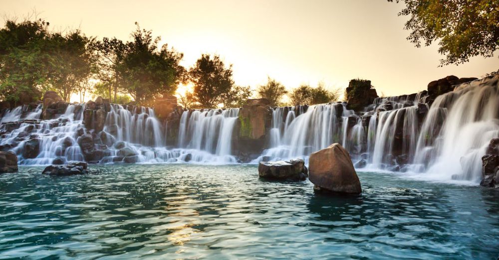 Summer Waterfalls - A serene waterfall surrounded by rocks and trees at sunset, radiating tranquility.