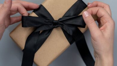 Gift Ideas - Hands tying a black ribbon on a craft paper gift box, next to scissors and twine.