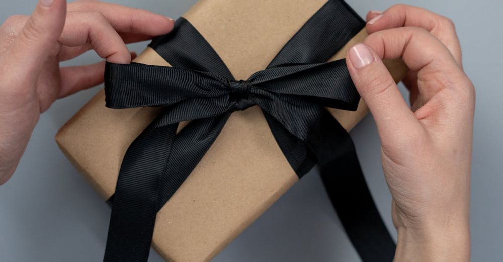 Gift Ideas - Hands tying a black ribbon on a craft paper gift box, next to scissors and twine.