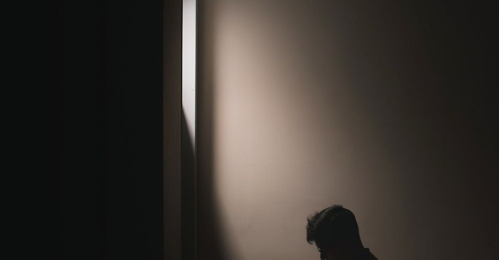 Books - Silhouette of a man sitting on a bed in a dark room, reading a book by window light.