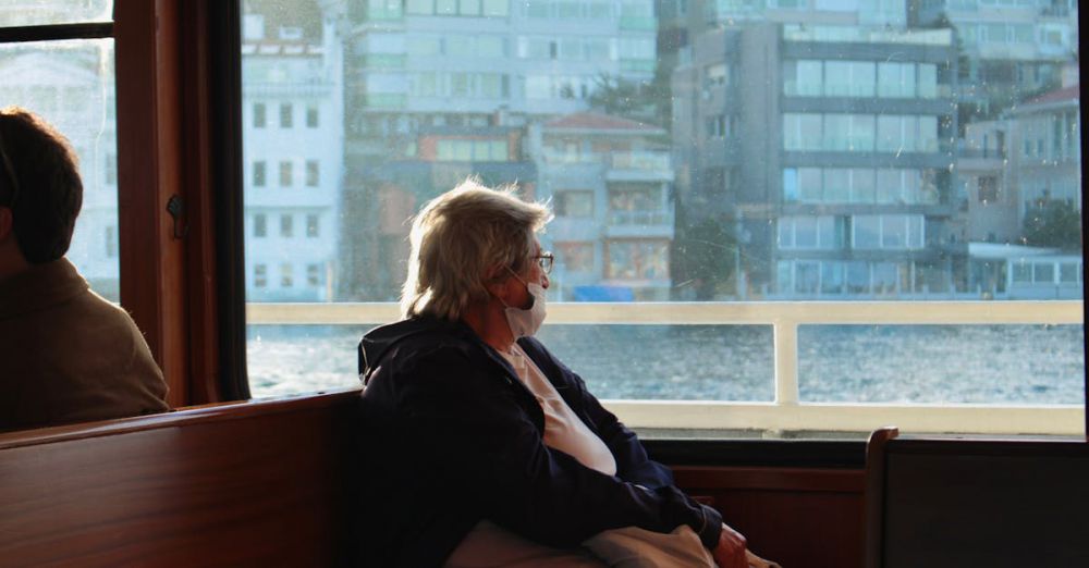 Portaging Safety - A senior woman wearing a mask gazes out the ferry window, reflecting by the sea.