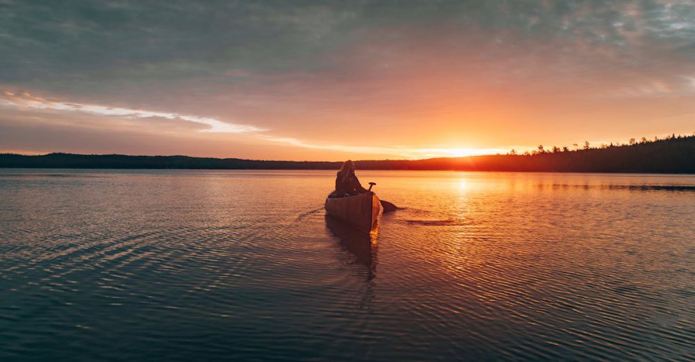 Solo Canoe Portaging - A lone canoeist paddling on Ely Lake at sunset, capturing nature's serene beauty.