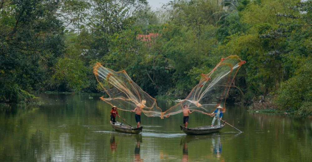 Multiday Canoe Portaging - Fishermen casting nets on Huong River near lush green foliage in Vietnam, capturing traditional lifestyle.