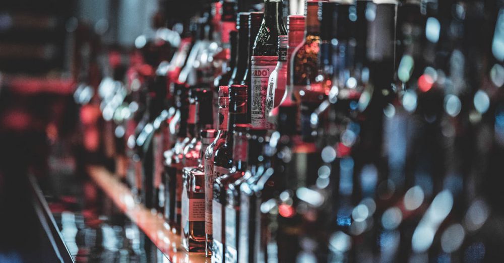 Wine & Beer Festivals - Blurred view of assorted alcohol bottles lined up on a bar shelf indoors.