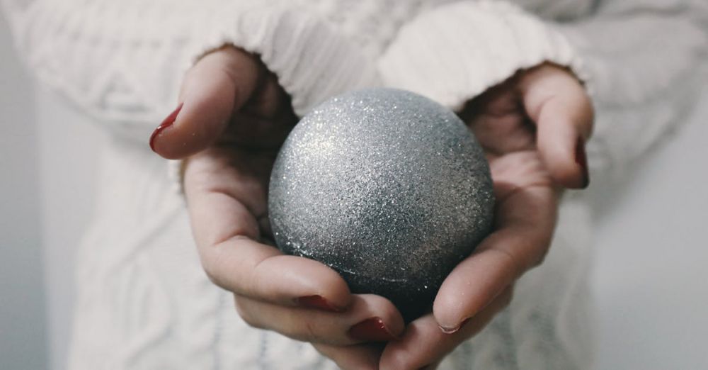 Artistic Winter Festivals - Close-up of female hands holding a glittery silver Christmas ornament in a cozy setting.