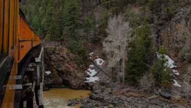 Rockies Scenic Train Rides - A train crossing a wooden bridge over a river in a lush mountain landscape during fall.