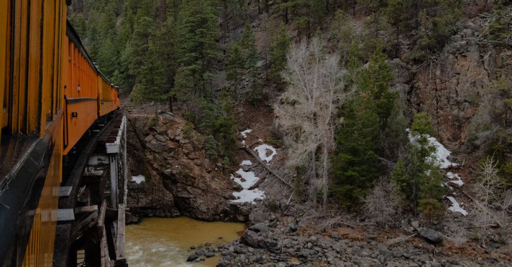 Rockies Scenic Train Rides - A train crossing a wooden bridge over a river in a lush mountain landscape during fall.