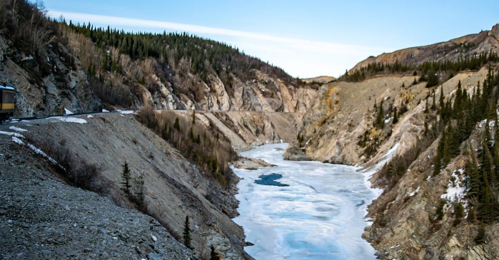 National Park Scenic Train Rides - Stunning view of frozen river cutting through rugged canyon in Alaska, under a clear blue sky.