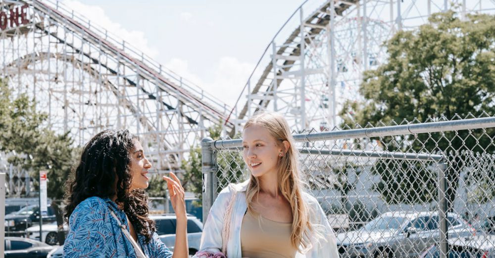 Weekend Scenic Train Rides - Two young women enjoying a sunny day at an amusement park, chatting happily near a rollercoaster.