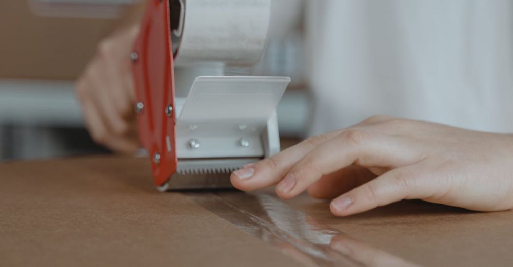 Tape Dispensers - A person sealing a cardboard box with a tape dispenser in an indoor setting.