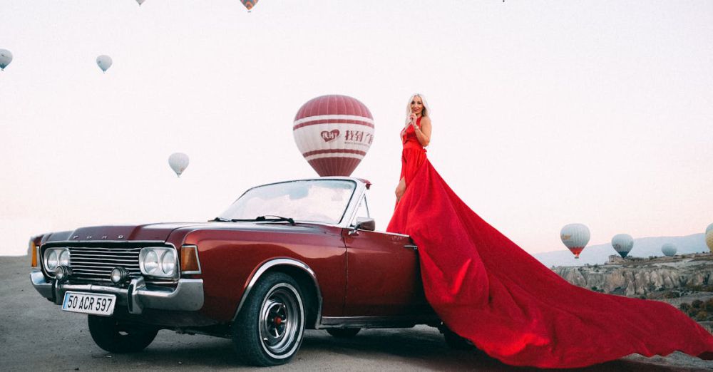 Adventure & Luxury Scenic Train Rides - Blonde woman in striking red dress posing by a vintage convertible with hot air balloons in the background.