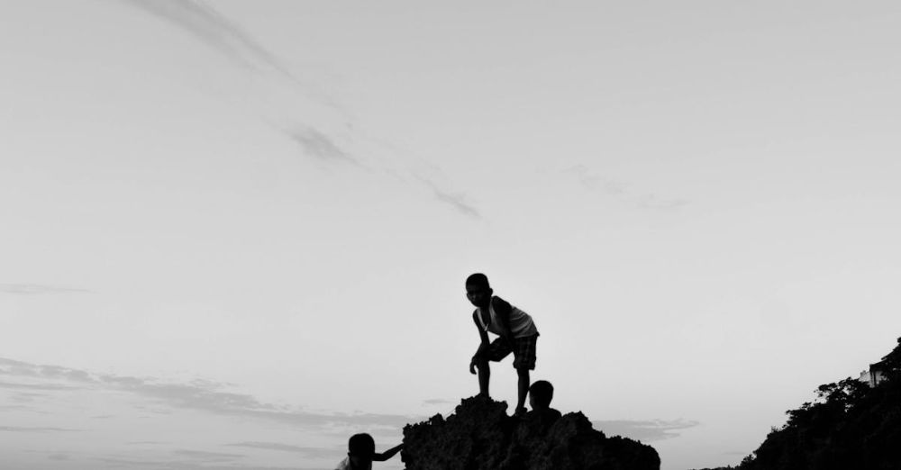 Family Rock Climbing - Black and white photo of children climbing rocks by the sea during twilight, creating a dramatic silhouette.