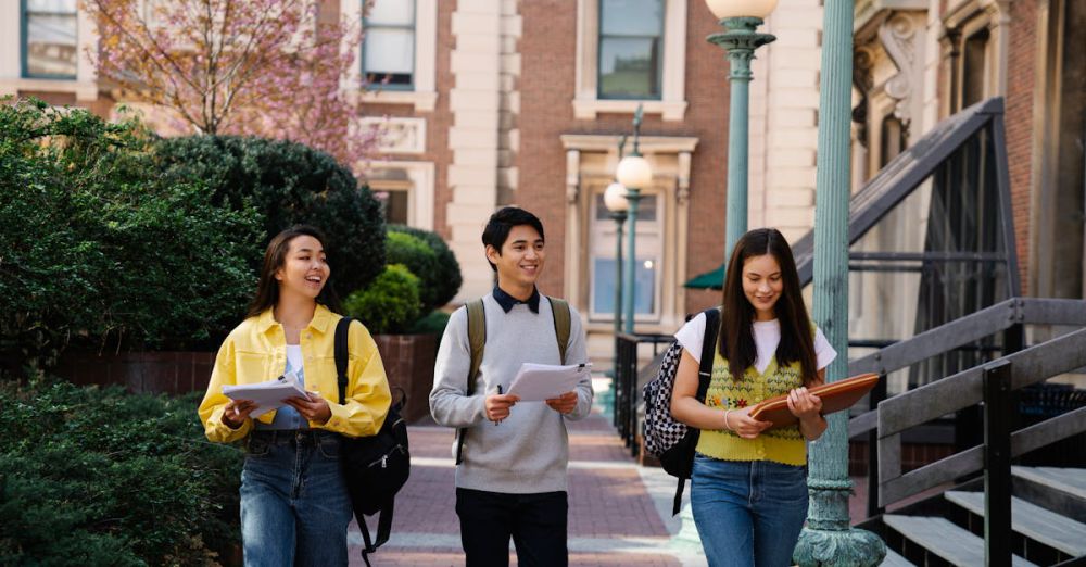 Interactive Urban Gardens - Three young adults walking and talking on a sunny campus day, holding notebooks and backpacks.