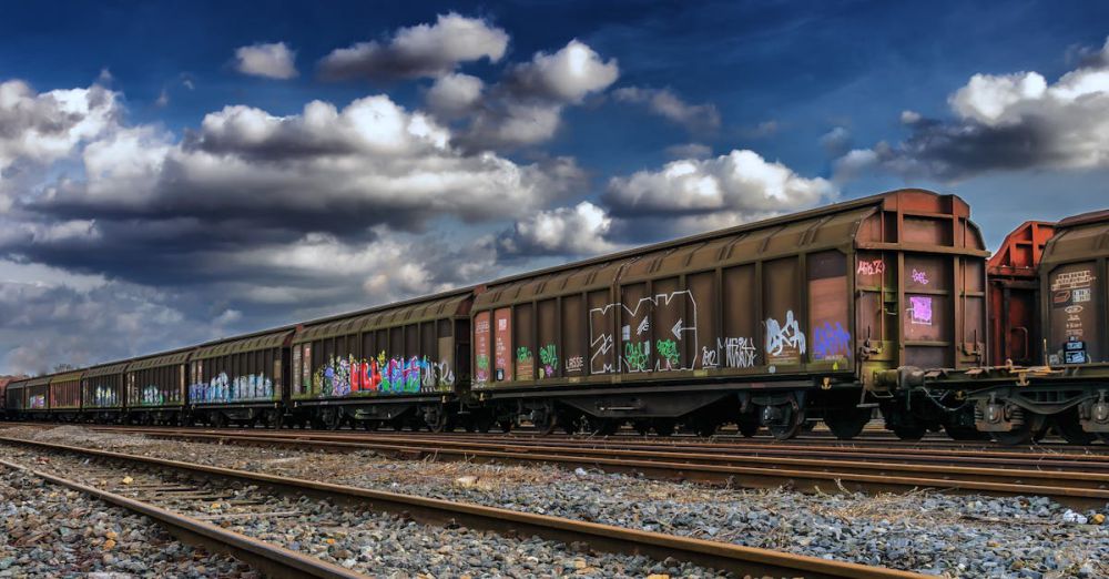 Goods - Freight train in a railway yard with colorful graffiti under a cloudy sky.