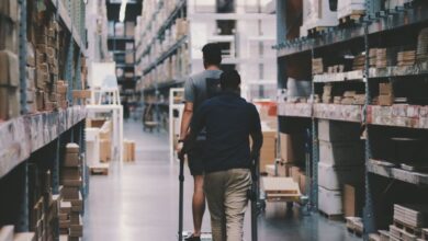 Goods - Two men maneuver a trolley in a large warehouse filled with boxes and shelves.