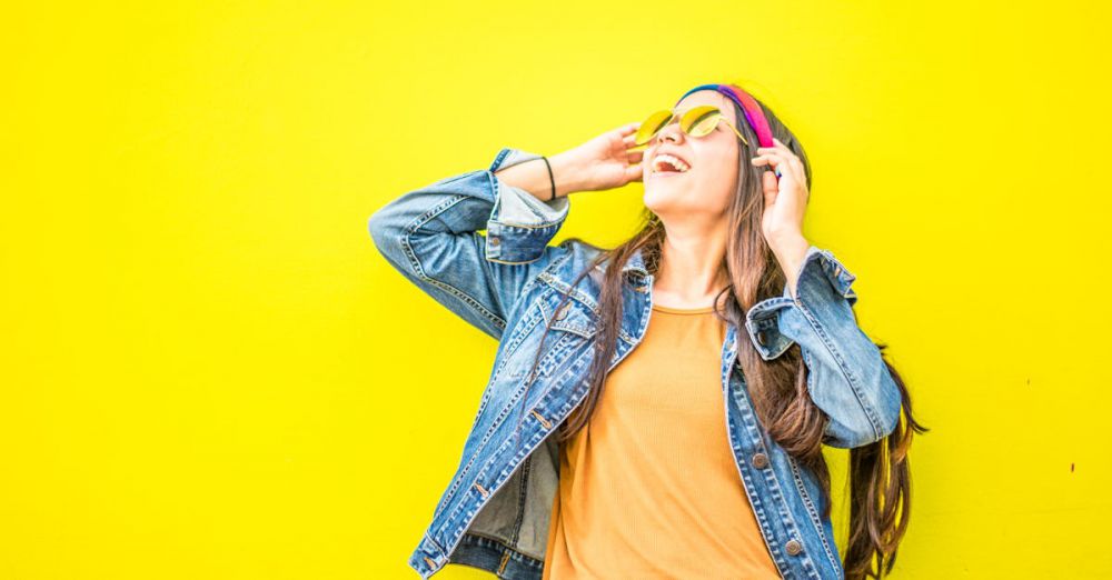 Fashion - Smiling woman in sunglasses stands against vibrant yellow wall, radiating happiness.