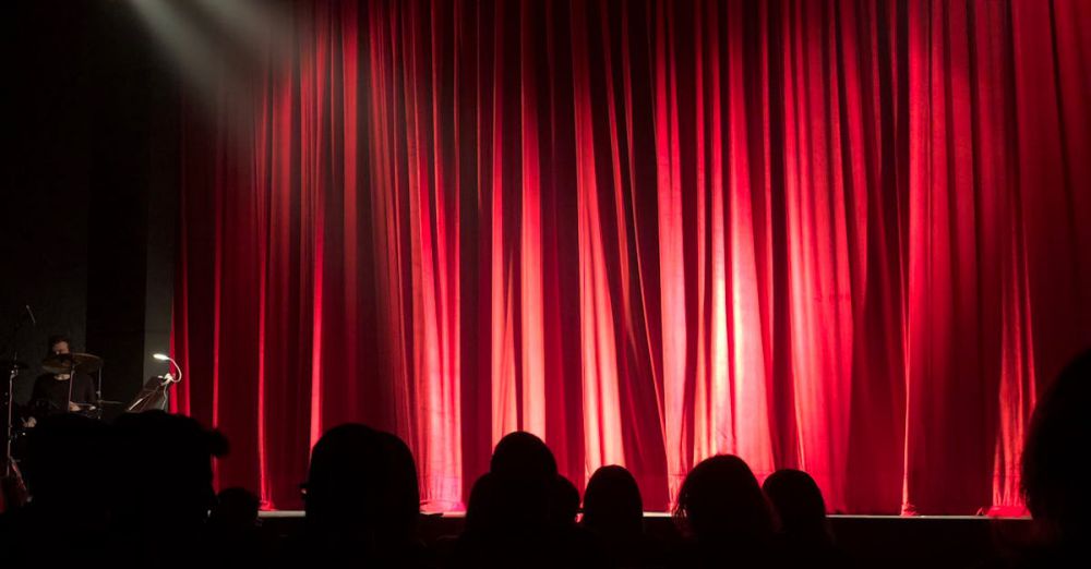 Theater - Dimly lit theater stage with red curtains and audience silhouettes under spotlights.