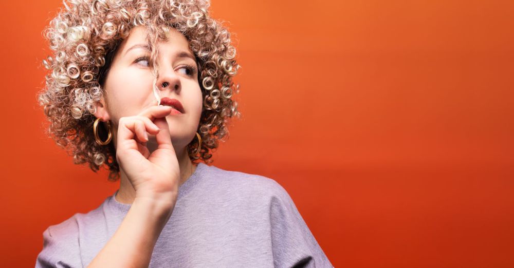 Comedy - Portrait of a young woman with curly hair, wearing a gray top, set against a vibrant red background.