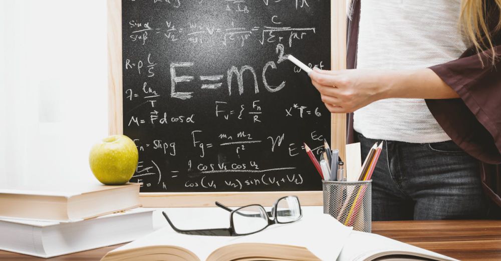 Lessons - Woman writing physics equations on a blackboard with books and an apple on the desk.