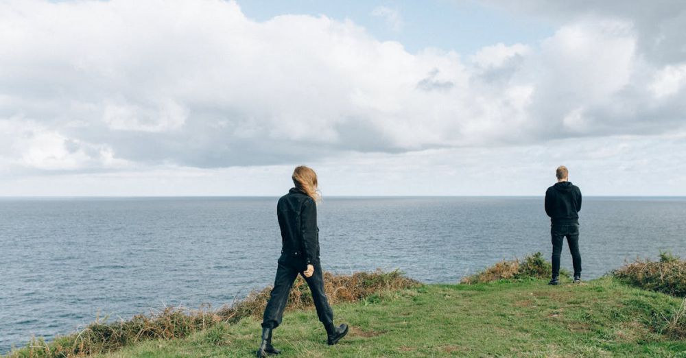 Hiking - Two people in dark clothing stand on a grassy cliff edge overlooking the ocean under a cloudy sky.