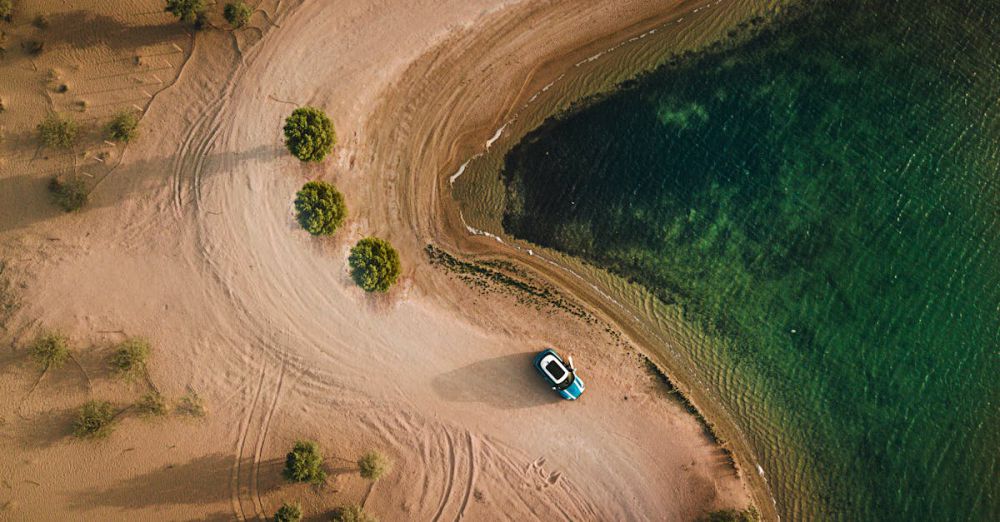 Trip - Aerial shot of a sandy beach, water, and parked car in Dubai.