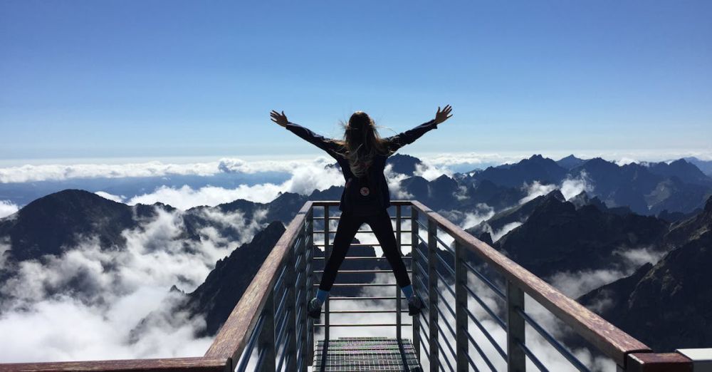 Adventures - A woman stands on a viewing platform in Vysoké Tatry, Slovakia, surrounded by clouds and mountains, embracing freedom.