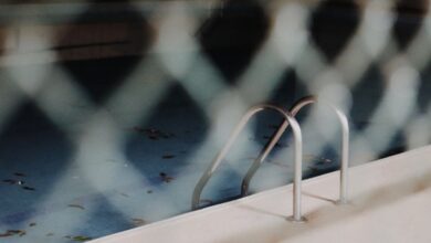 Off-Season - A view of a neglected swimming pool with a metal ladder, seen through a chain-link fence.