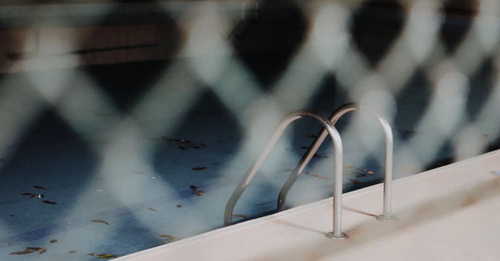 Off-Season - A view of a neglected swimming pool with a metal ladder, seen through a chain-link fence.