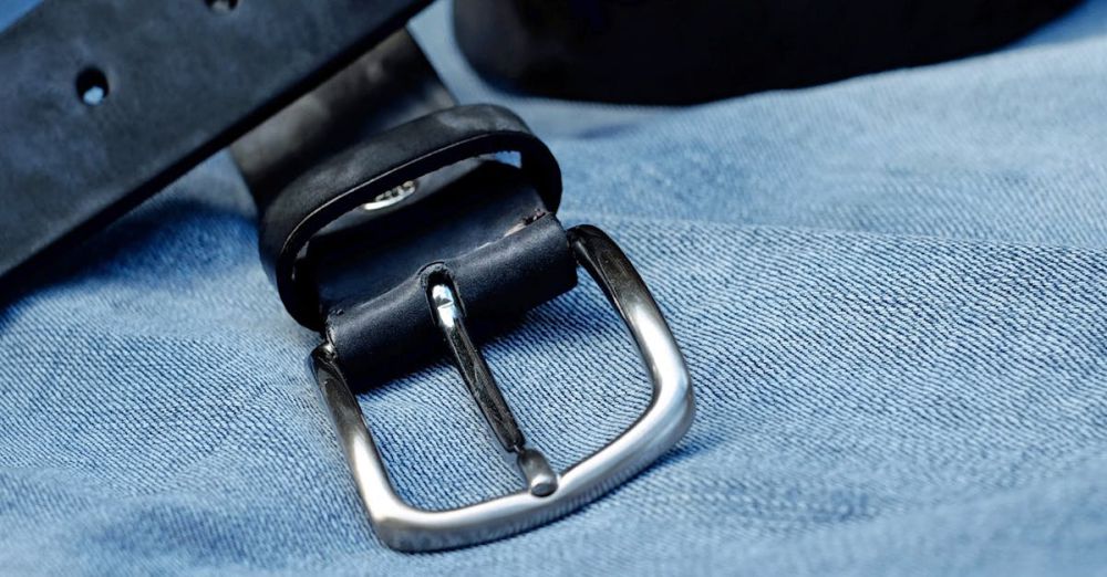 Belts - Close-up of a black leather belt with metal buckle laid on blue denim fabric.