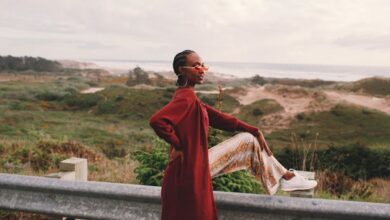 Sunglasses - Side view of young stylish African American female traveler in trendy outfit and sunglasses sitting on road metal fence during trip near ocean against cloudy sky