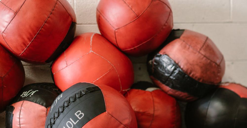 Weighted Balls - Red and black medicine balls stacked in a gym setting with two blue hydration cartons on a dark floor.