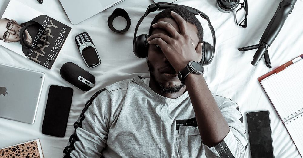 Gadgets - A young man lying on a bed surrounded by modern gadgets, books, and tech devices, illustrating digital lifestyle.