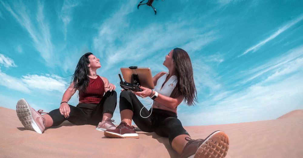 Gadgets - Two women on a Dubai sand dune control a drone under a bright blue sky.