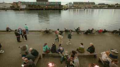 Fire Pits - A bustling riverside promenade with people enjoying outdoor fire pits and a serene water view.