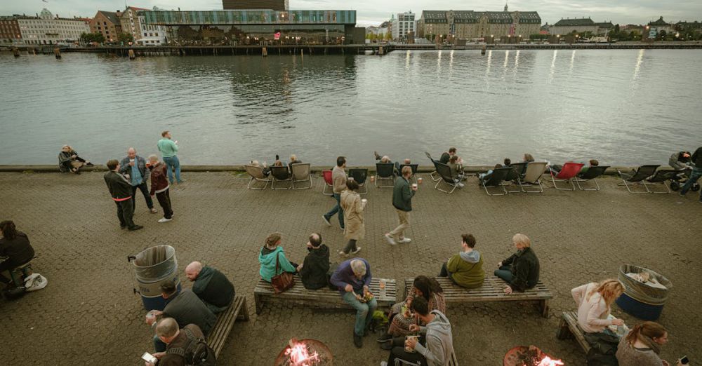 Fire Pits - A bustling riverside promenade with people enjoying outdoor fire pits and a serene water view.