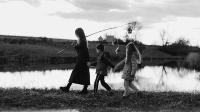 Birdhouses - A mother and children walk by a tranquil lake, carrying birdhouses on a stick, in black and white.