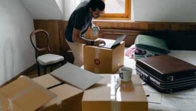 Fitness Boxes - Side view of ethnic male in casual clothes and wristwatch surfing internet on netbook and standing with cup of hot drink surrounded by cardboard boxes and suitcases in bedroom