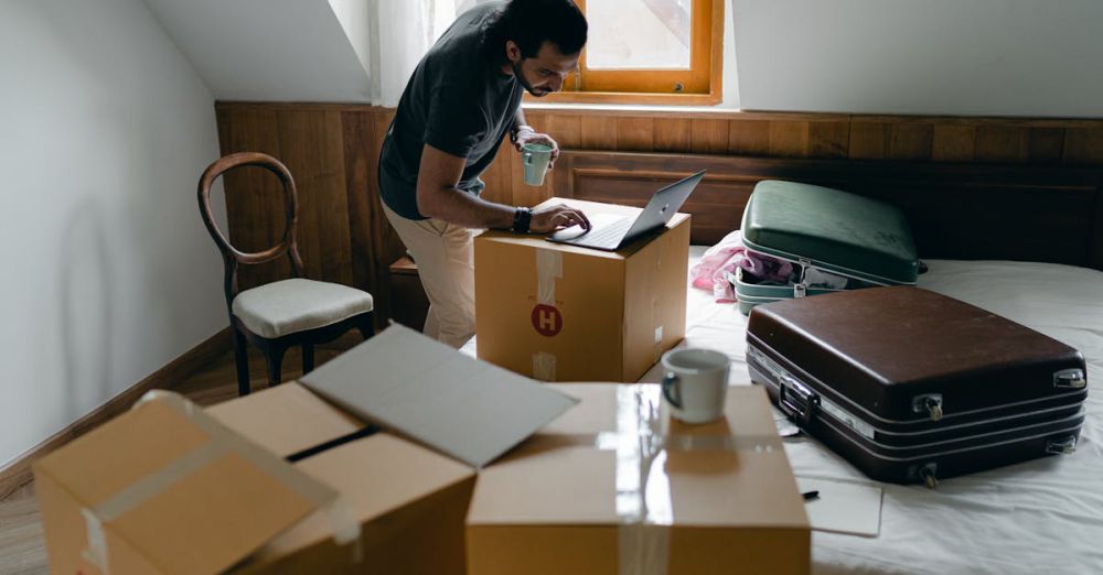 Fitness Boxes - Side view of ethnic male in casual clothes and wristwatch surfing internet on netbook and standing with cup of hot drink surrounded by cardboard boxes and suitcases in bedroom