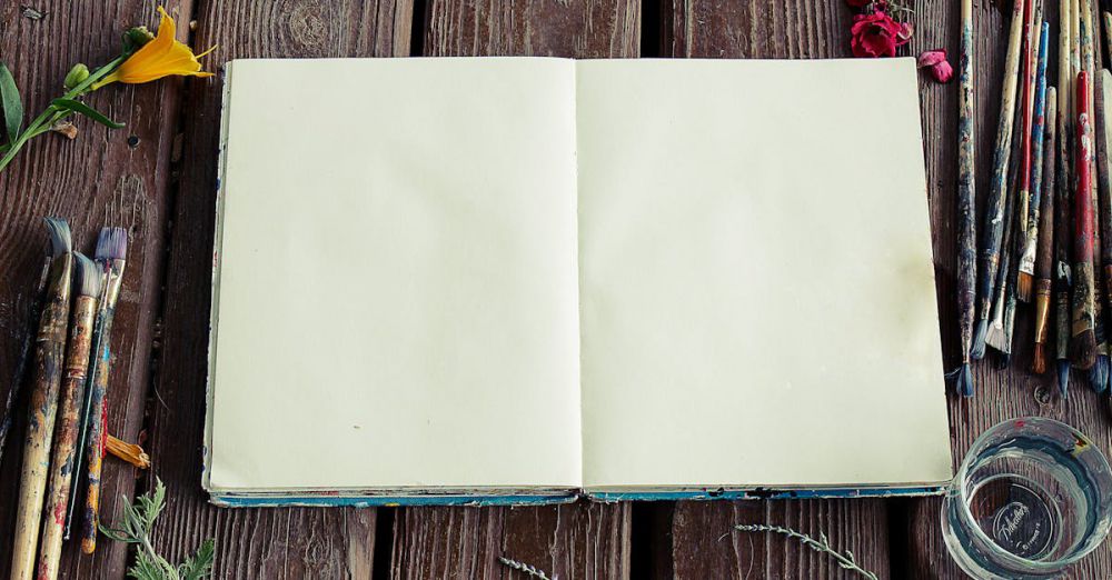 Art Supplies - Blank sketchbook surrounded by paintbrushes and flowers on rustic wooden surface.