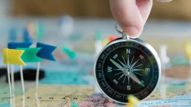 Geography Tools - Close-up of hand holding compass over a map with colorful pins and flags, symbolizing travel planning.