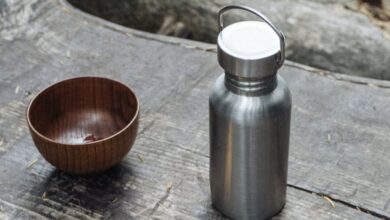 Camping Utensils - A wooden bowl, fork, and reusable stainless steel flask on a wooden table outdoors, emphasizing sustainability.