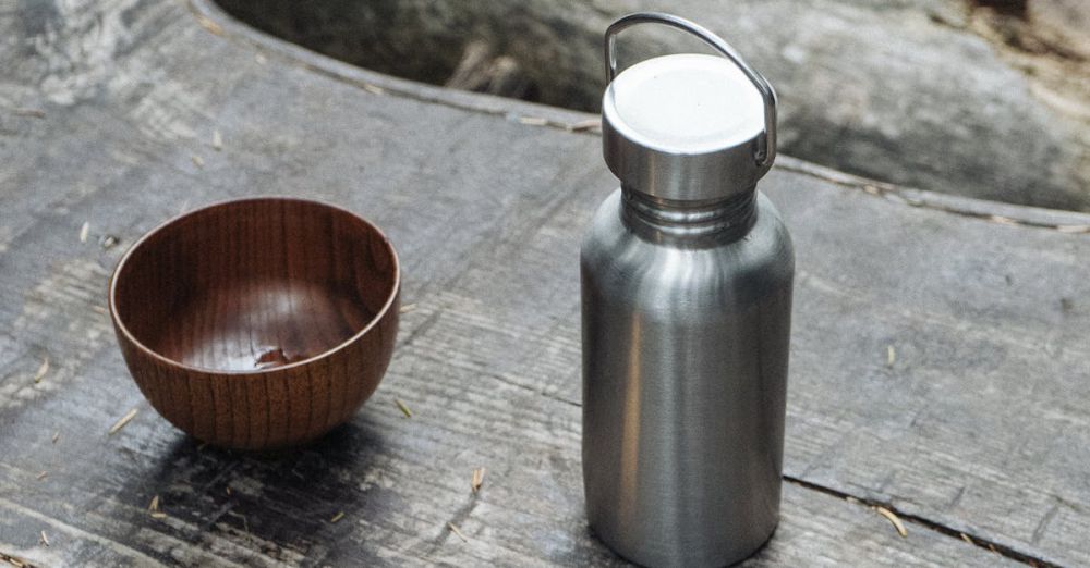 Camping Utensils - A wooden bowl, fork, and reusable stainless steel flask on a wooden table outdoors, emphasizing sustainability.