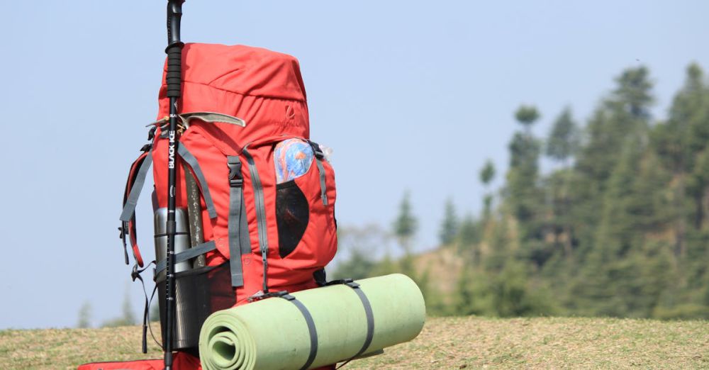 Camp Mats - Red backpack with hiking pole and mat on grassy field with forest background.