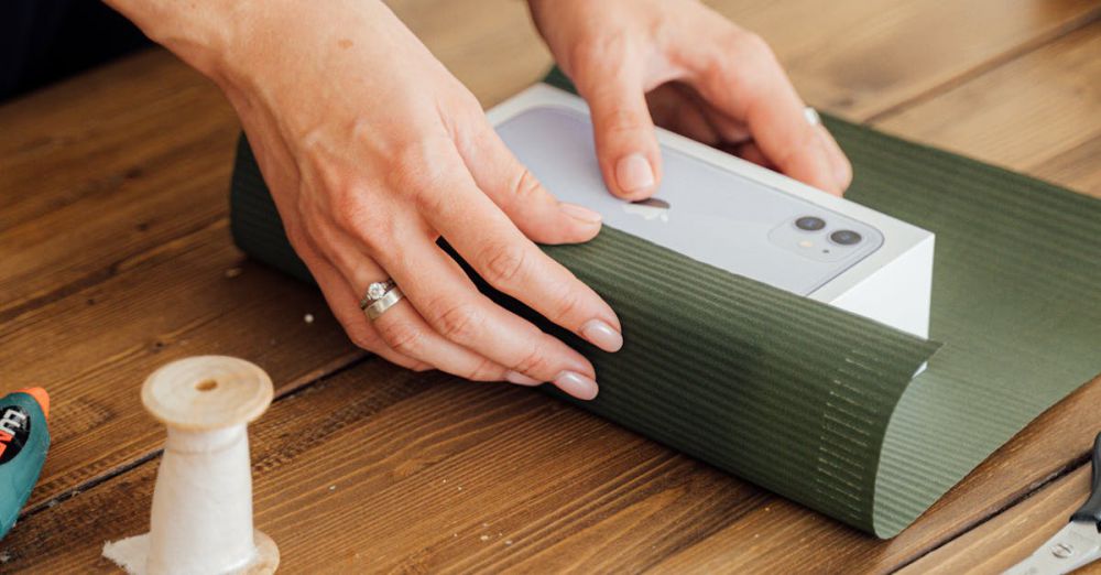 Gift - Close-up of hands wrapping a gift box with green paper on a wooden table, surrounded by sewing supplies.